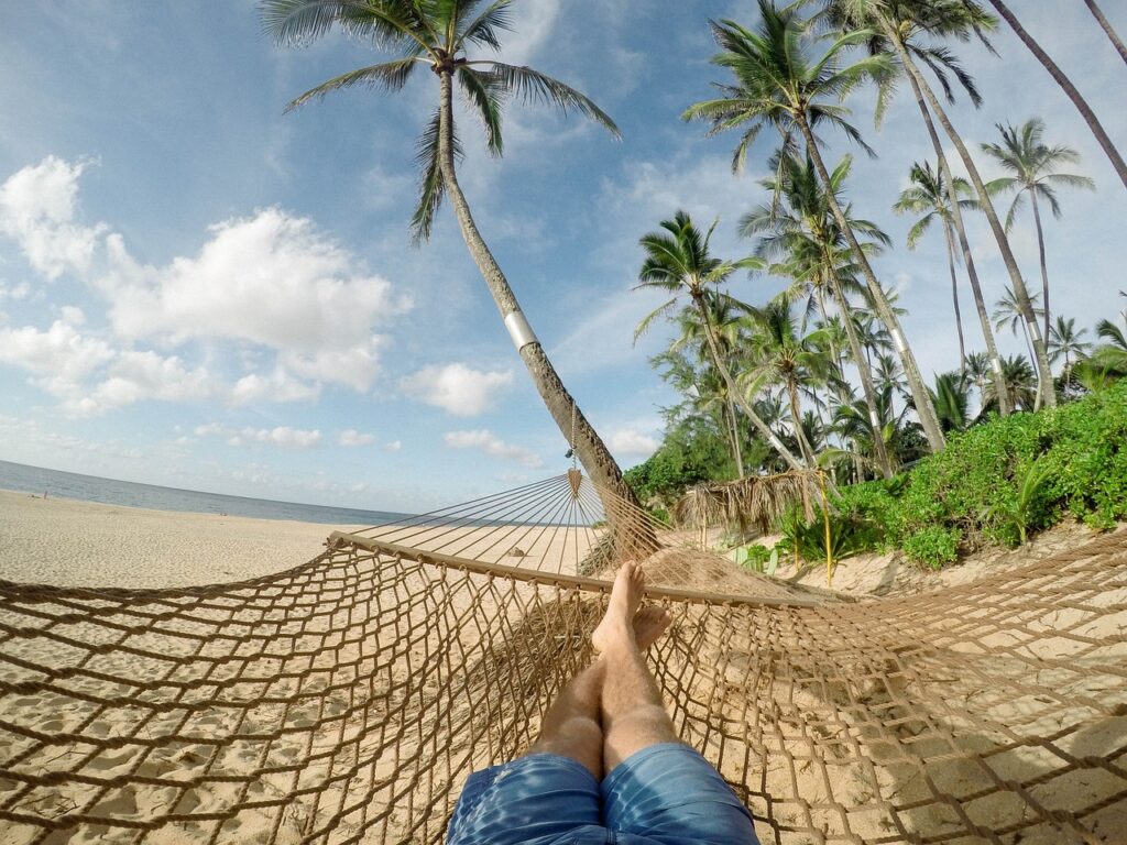 Photo of a hammock on a beach with palm trees and a person's legs and feet lying on the hammock.